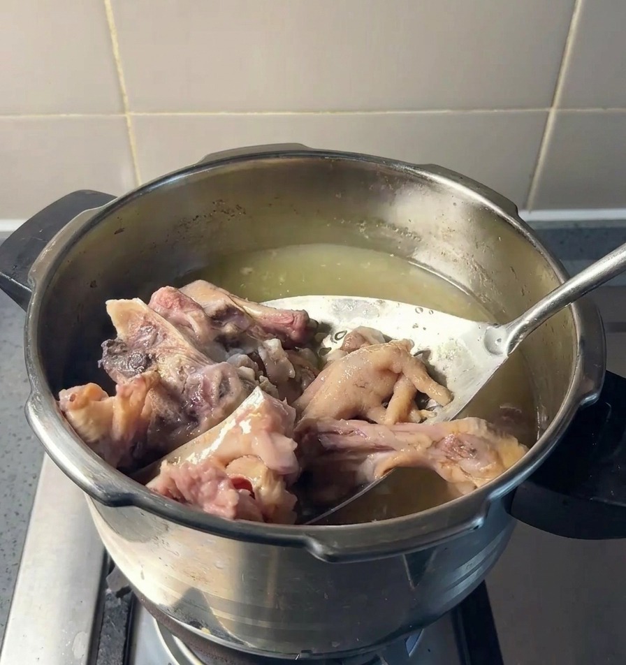 A stainless steel slotted strainer lifting large pieces of cooked pork bone and chicken feet out of a simmering pot of clear yellowish broth.