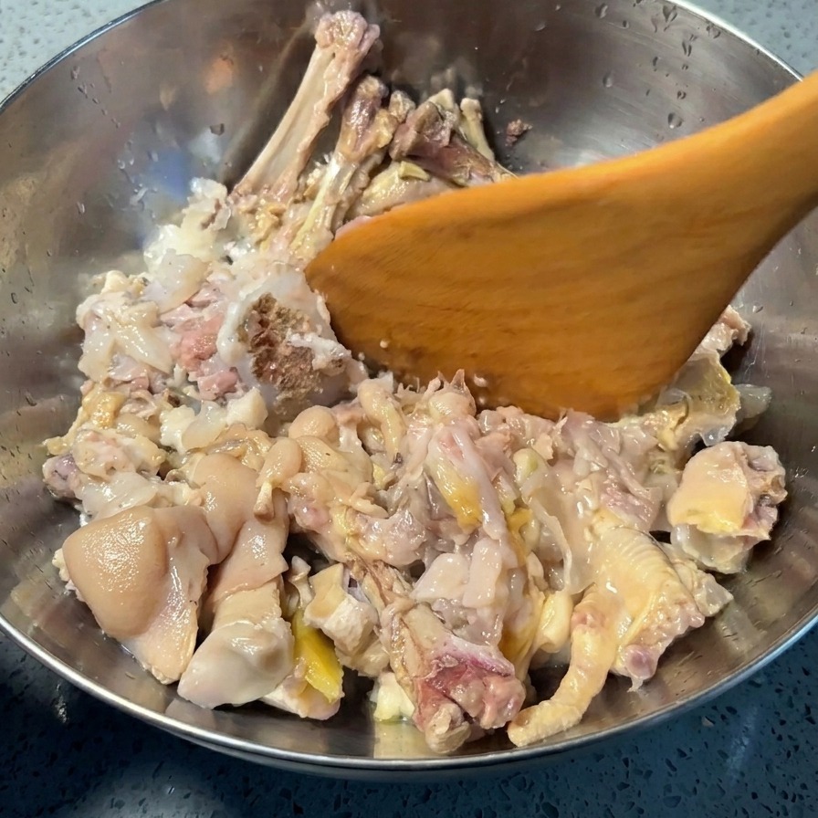 A wooden spatula pressing down on cooked pigs trotter and chicken feet inside a silver mixing bowl to separate meat and skin from the bones.