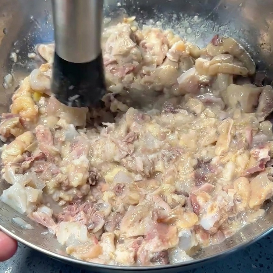 A metal hand masher crushing cooked pork and chicken meat into a chunky paste at the bottom of a stainless steel bowl.