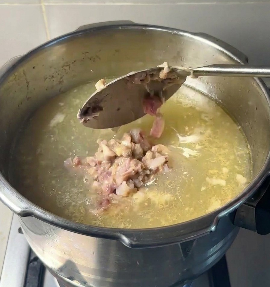A large metal spoon dropping clumps of mashed cooked meat and marrow into a pot of clear simmering soup base.