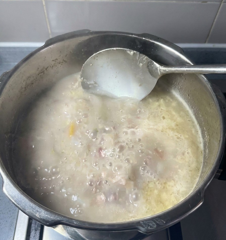 A large metal ladle stirring a pot of opaque, milky-white pork bone broth with visible bits of tender meat and fat swirling in the liquid.