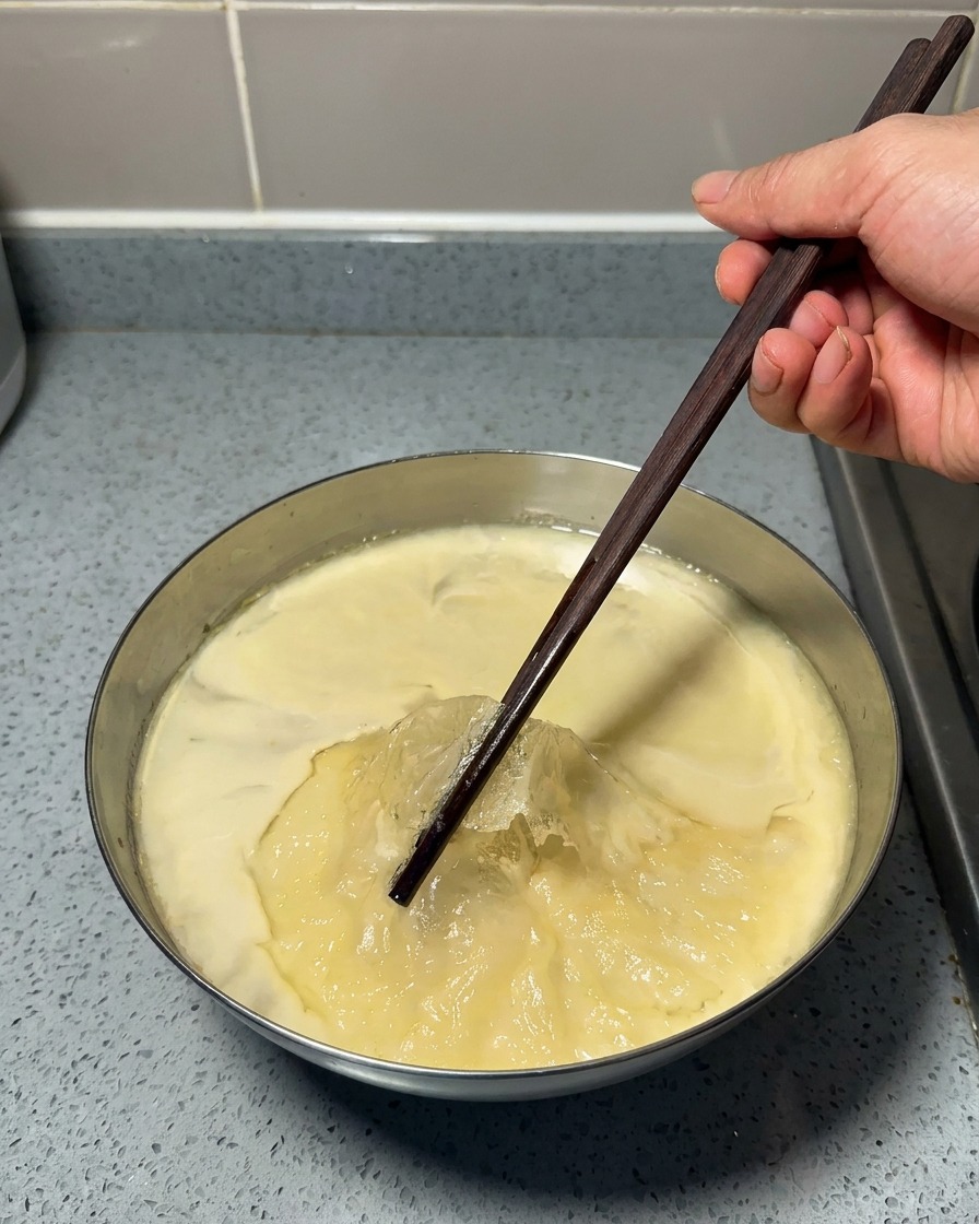 A hand using dark wooden chopsticks to peel a thin layer of solidified yellow fat off the top of a chilled, jelly-like soup base in a metal bowl.