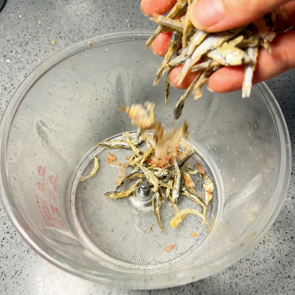 A hand pouring dried anchovies and bonito flakes into a clear kitchen blender jar.