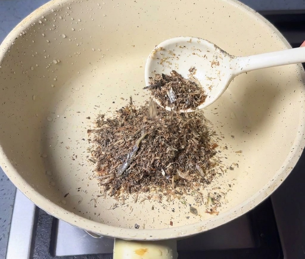 A spoonful of fine brown seafood powder being added to a dry, light-colored saucepan.