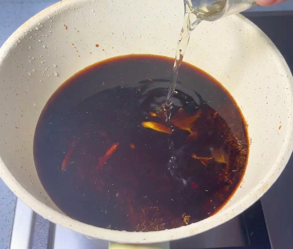 Clear liquid being poured from a glass into a beige pan containing dark liquid and ground fish powder.