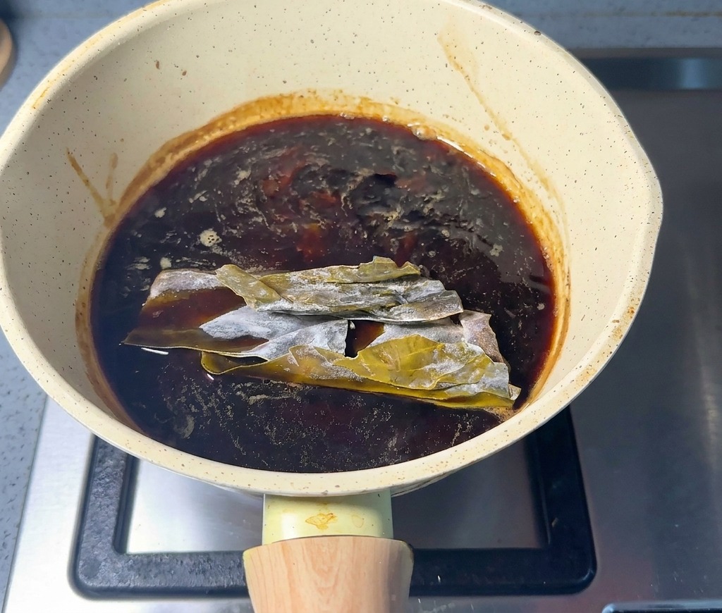 Slabs of dried green kombu submerged in the dark seasoning sauce inside a light-colored saucepan.