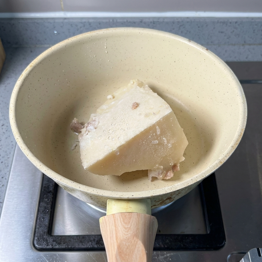 A block of solid frozen white pork bone broth and a spoonful of fish powder in a beige saucepan.
