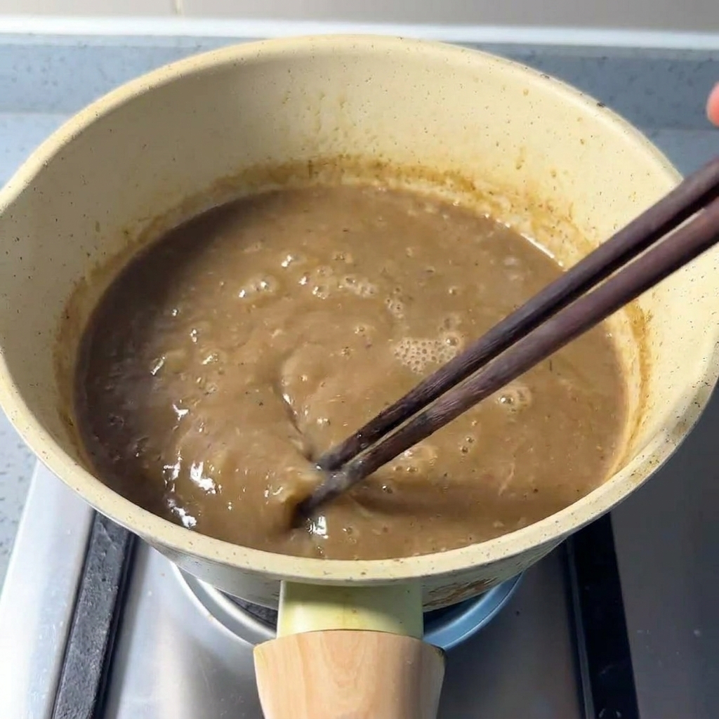 Stirring a thick, brownish pork bone broth mixed with seafood powder in a pan using chopsticks.