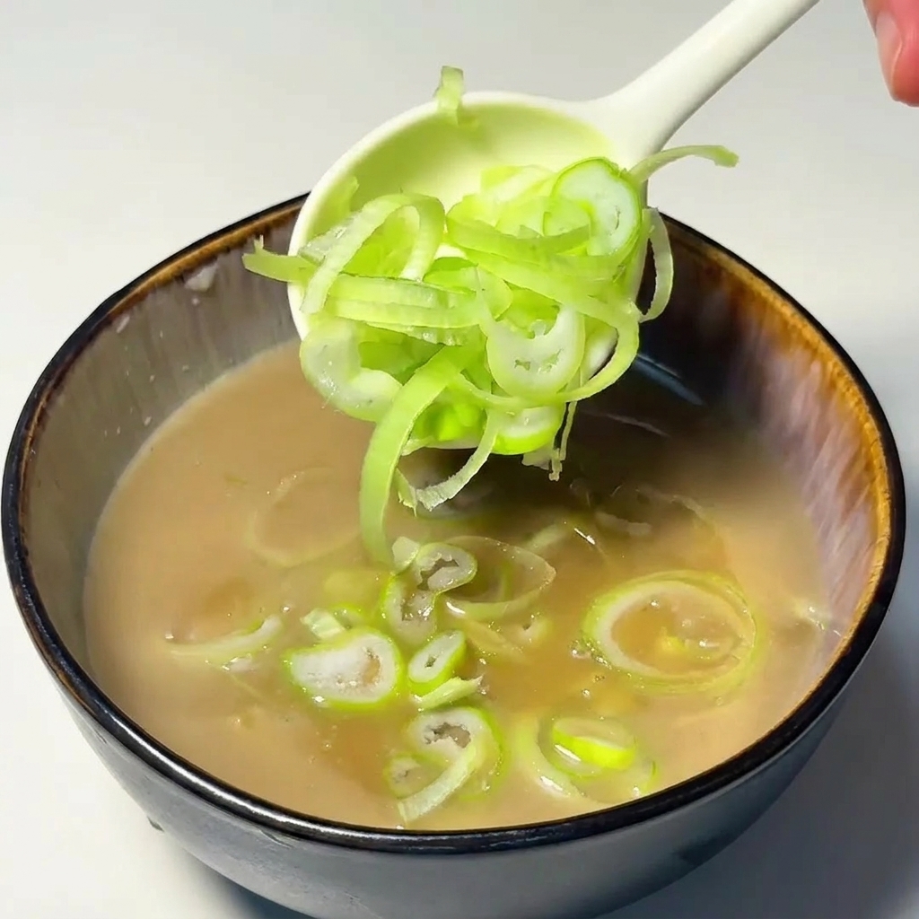 A white spoon depositing a pile of thinly sliced green onion rings into the tan-colored dipping soup.
