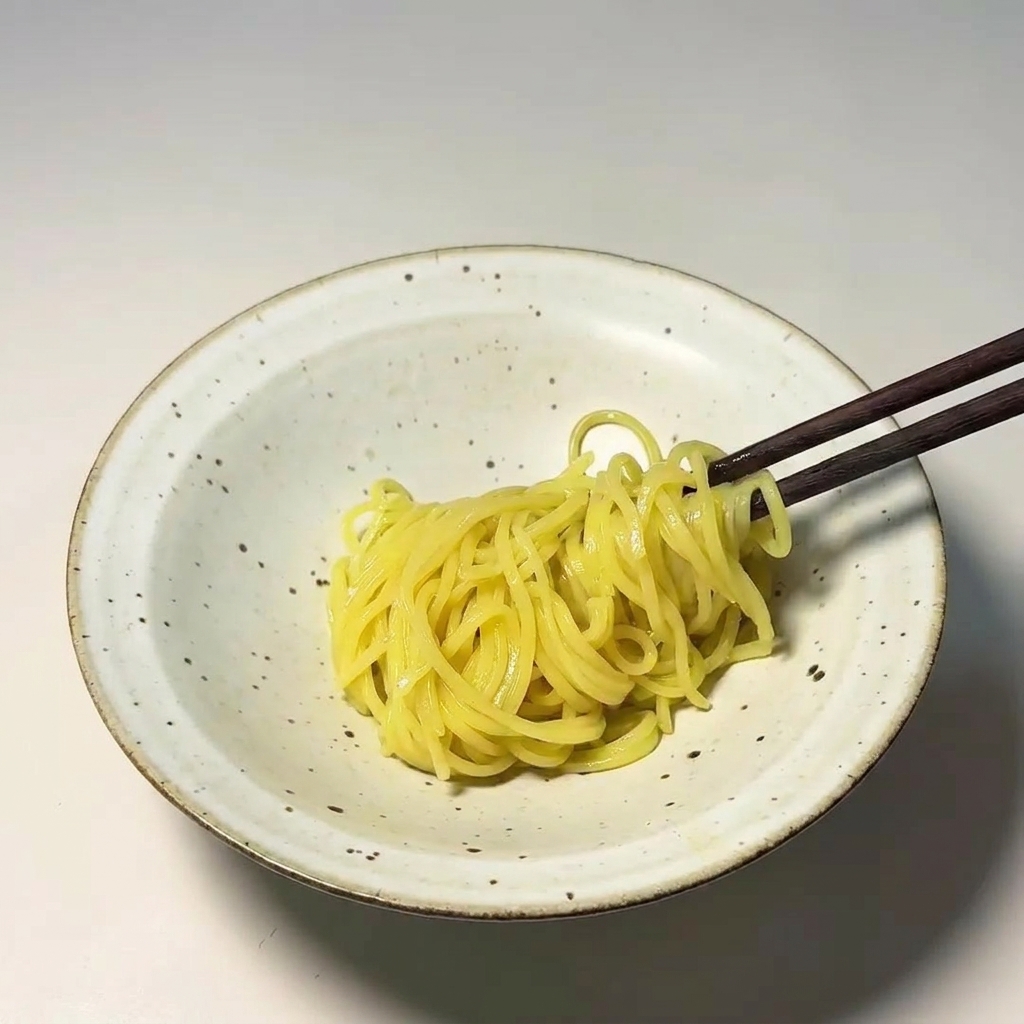 Dark wooden chopsticks placing a bundle of cooked yellow noodles into a speckled white ceramic bowl.