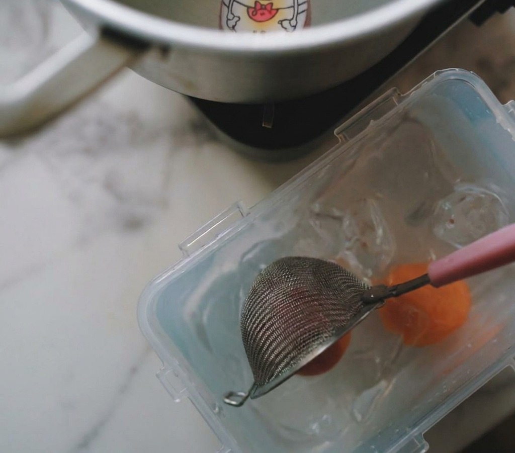 A mesh strainer lifting a boiled egg from a pot and placing it into a clear container filled with ice and water.