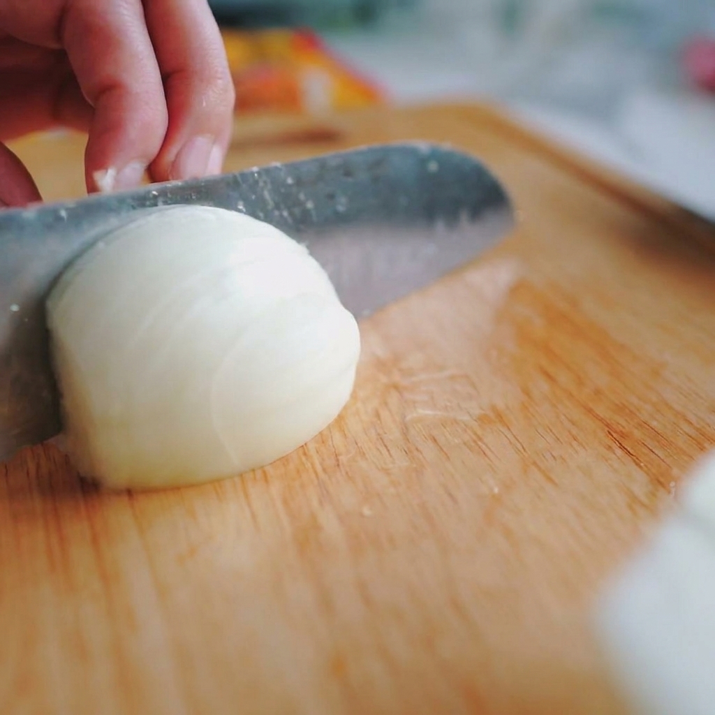 A close-up shot of a hand holding a white onion and slicing it into thin vertical strips with a chefs knife on a wooden board.