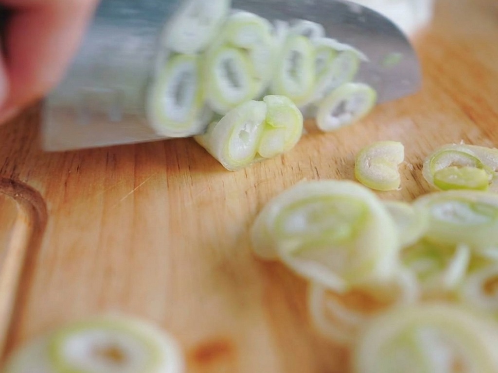 Close-up of hands using a sharp chefs knife to slice stalks of green onions into thin ring-shaped cross-sections on a wooden cutting board.