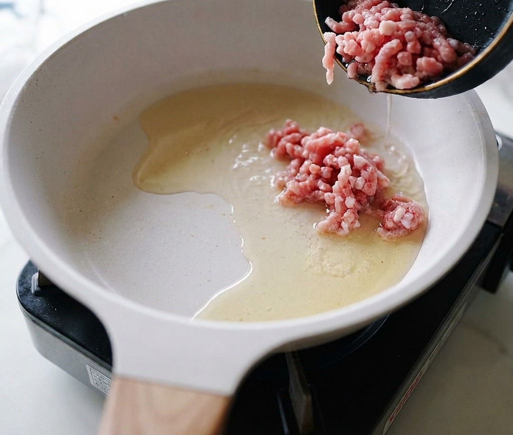 Raw ground pork being poured from a small black skillet into a white ceramic pan containing heated cooking oil.