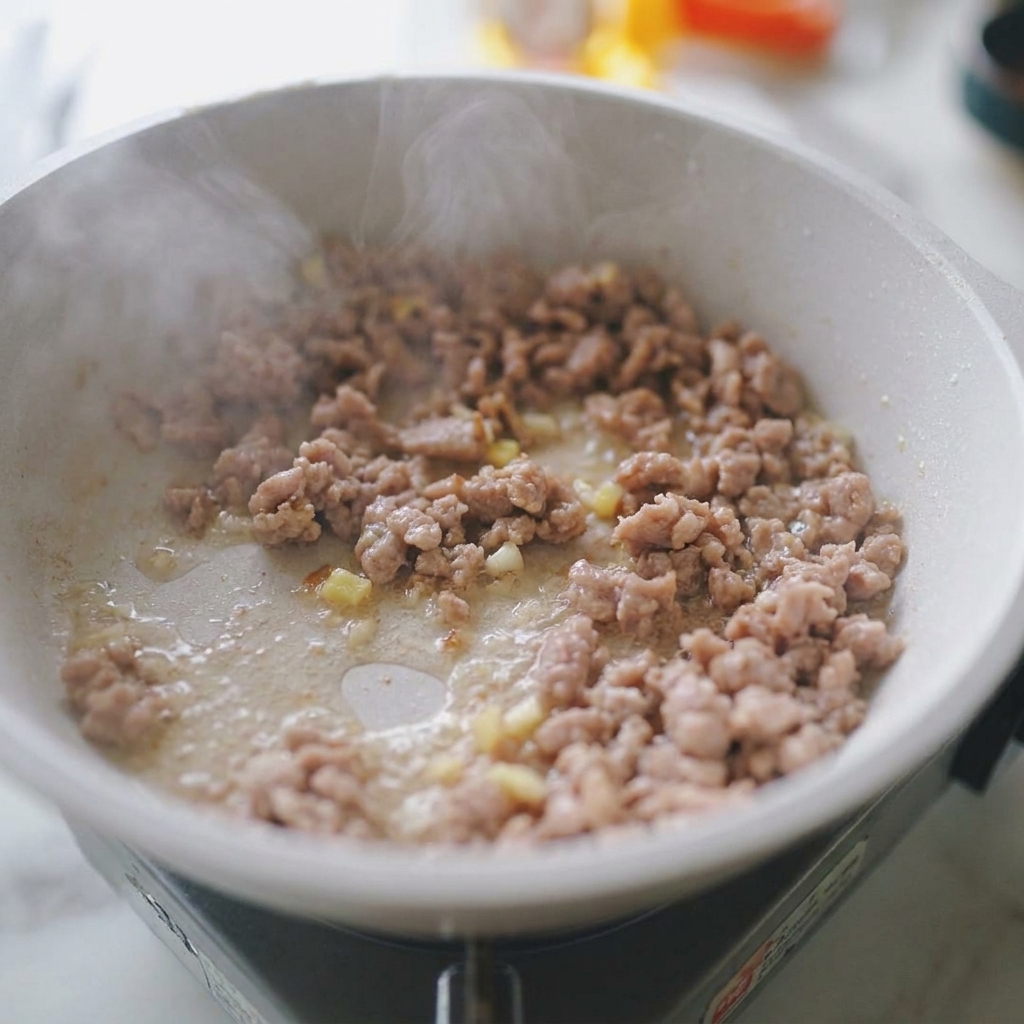 Minced pork being stir-fried in a white pan with visible pieces of minced garlic and ginger, with steam rising from the meat.