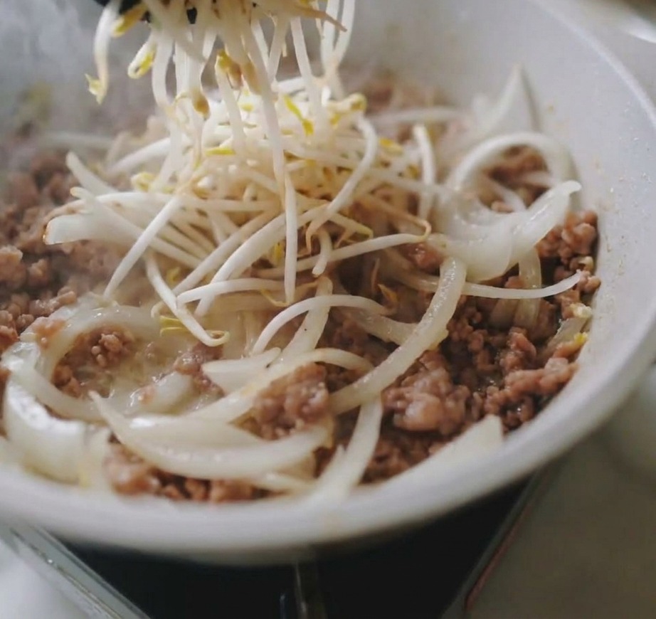 A hand pouring a large pile of fresh white bean sprouts into a skillet containing cooked minced pork and translucent onion slicescite: 1.