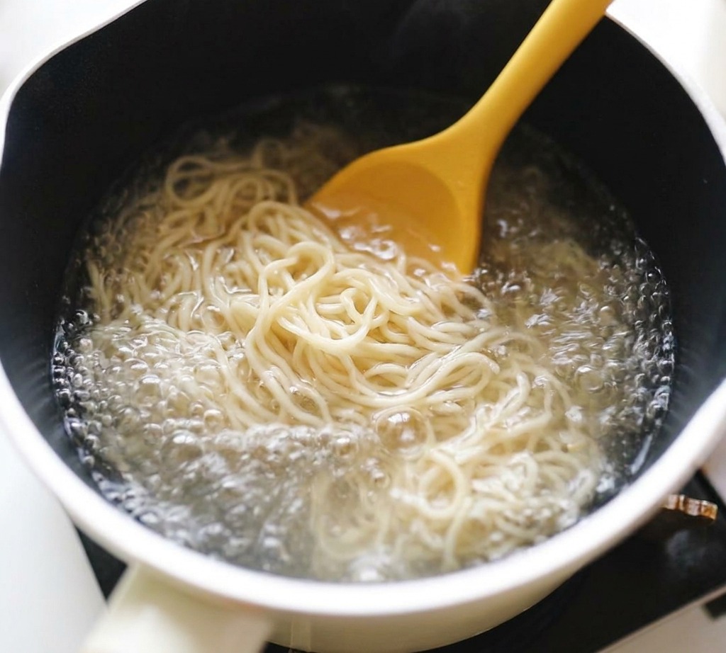 Wavy ramen noodles being stirred with a bright yellow spoon in a white pot of vigorously bubbling watercite: 1.