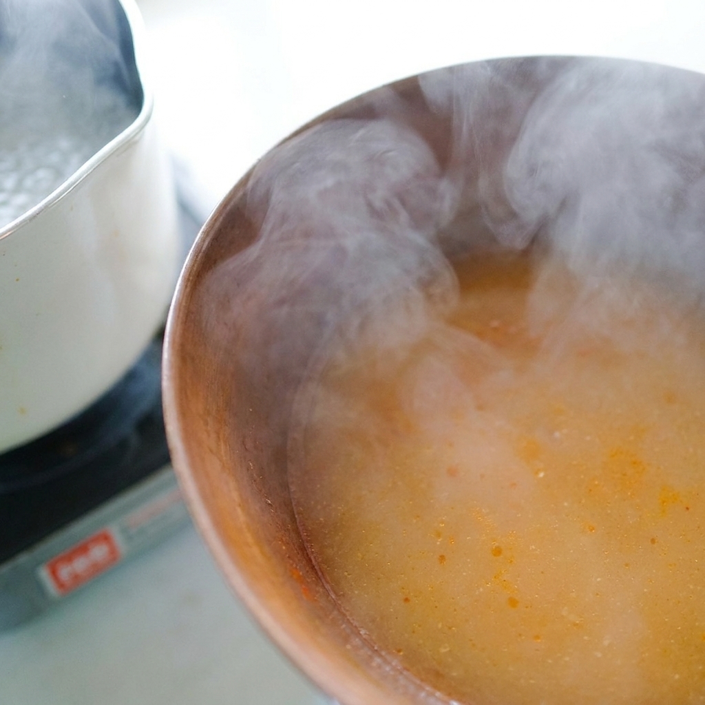 Hot, steaming noodle water being poured from a ladle into a bowl with miso paste to form a broth.