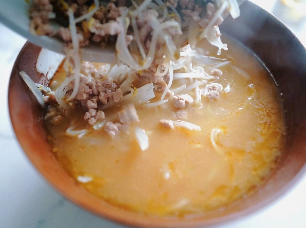 A close-up view of a metal spoon transferring stir-fried minced pork and bean sprouts into a wooden bowl containing ramen noodles and orange-colored miso broth.