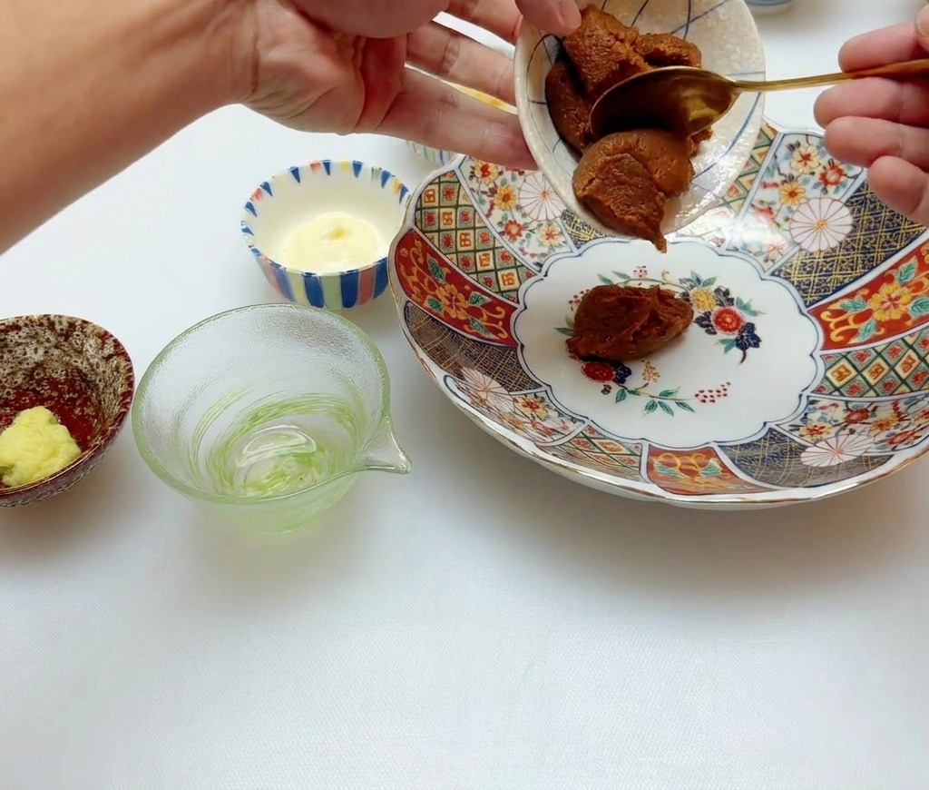 A hand scooping thick brown miso paste from a small ceramic dish into a larger floral patterned bowl using a gold spoon.