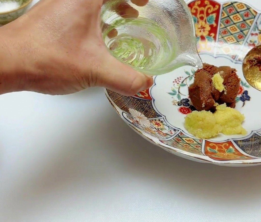 Clear salad oil being poured from a small green-tinted glass pitcher onto miso and ginger paste in a bowl.