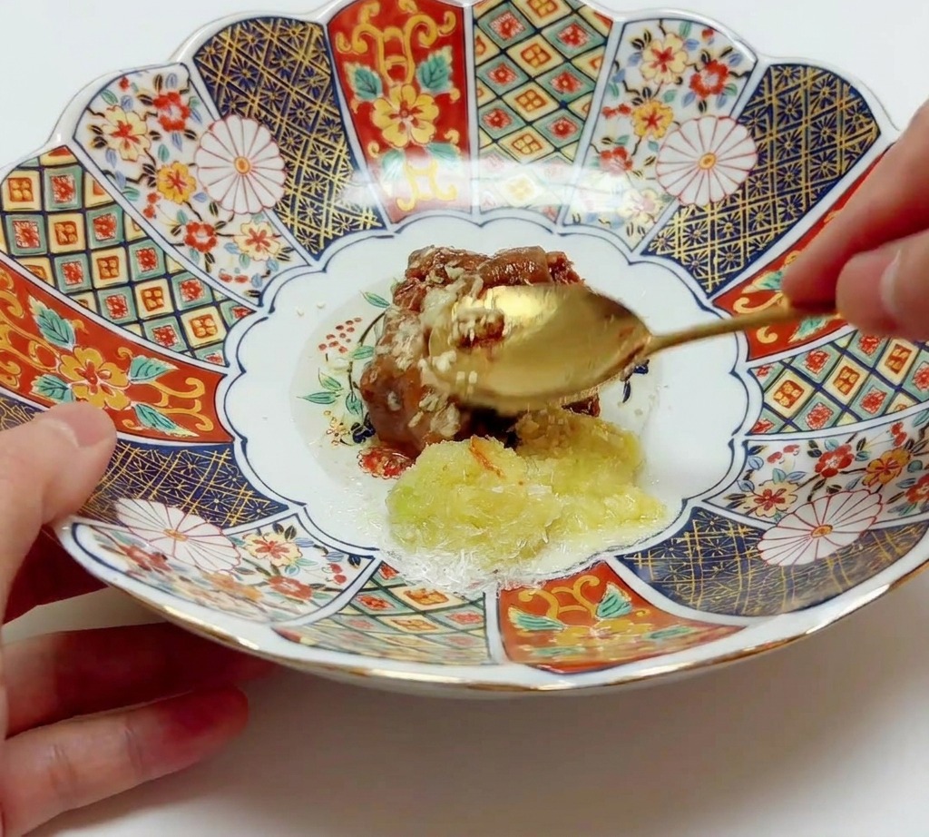 A gold spoon stirring and combining brown miso paste with aromatics and oil in a large patterned ceramic bowl.