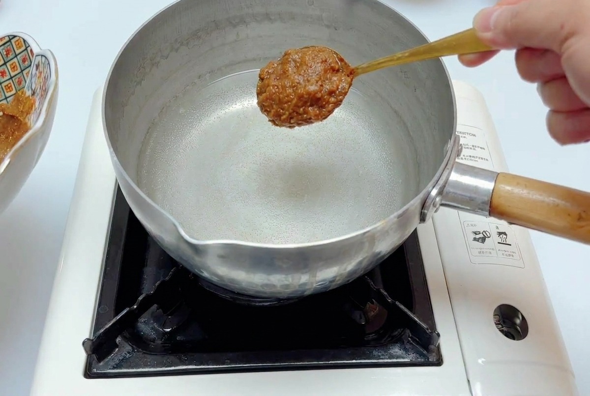 A spoonful of thick miso seasoning sauce held over a stainless steel pot of clear water on a portable stove.