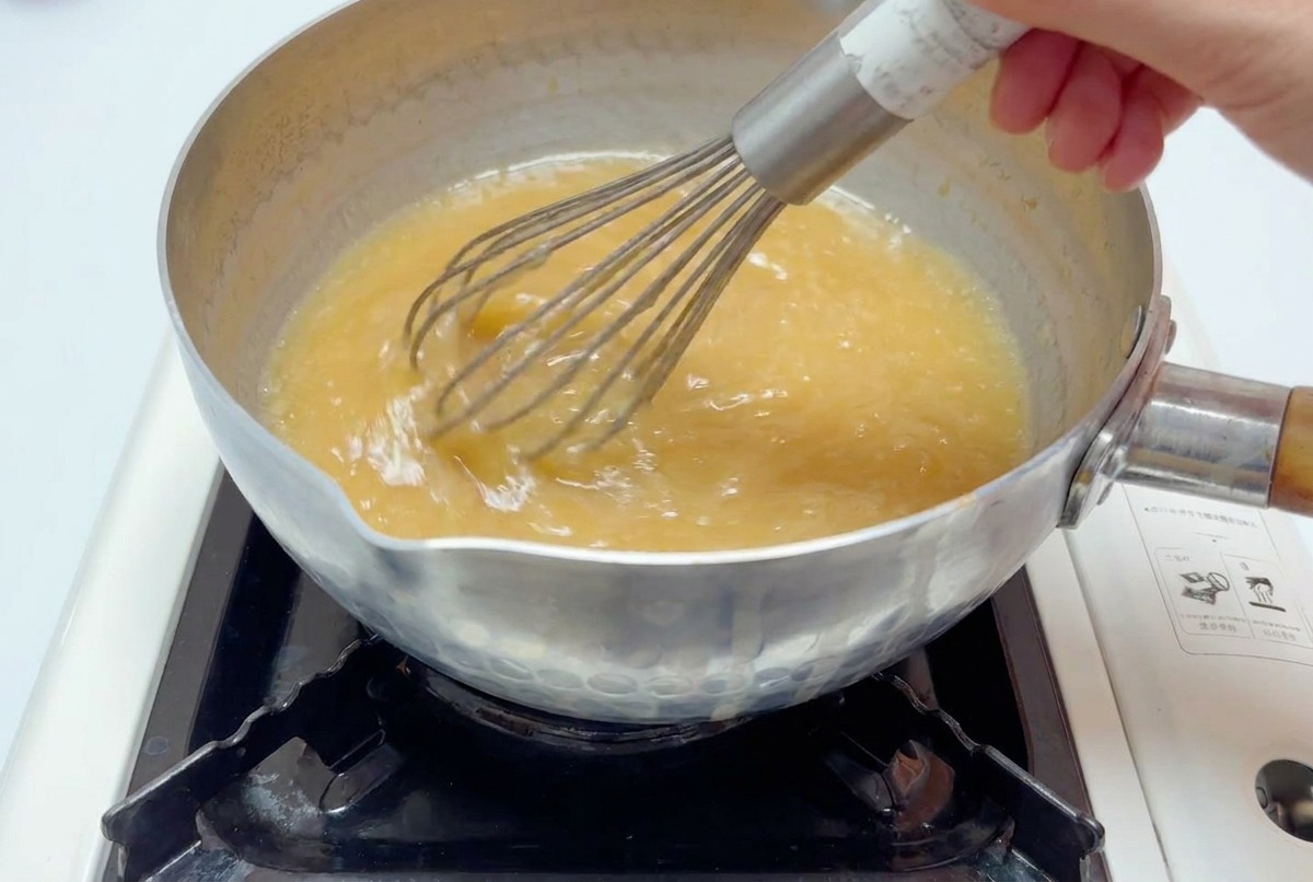 A metal whisk stirring a light brown miso broth in a pot over a flame until it is fully blended.