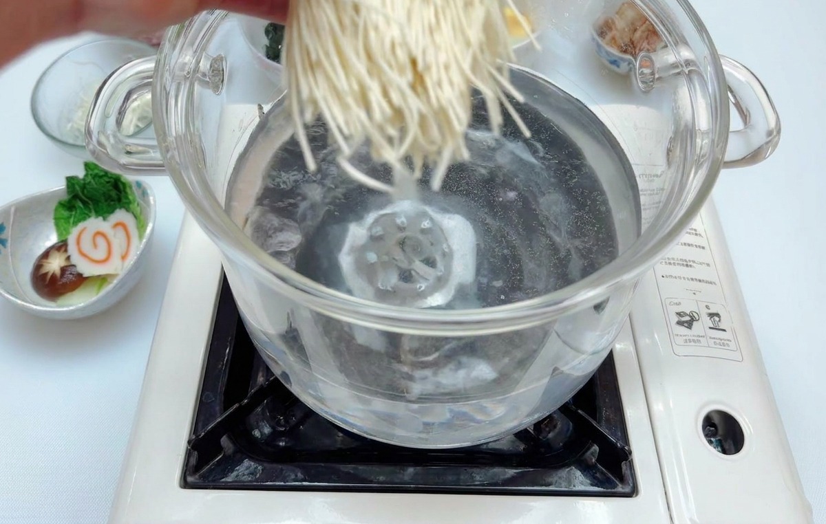 A hand dropping a bundle of thin white ramen noodles into a glass pot of clear boiling water on a portable induction cooktop.