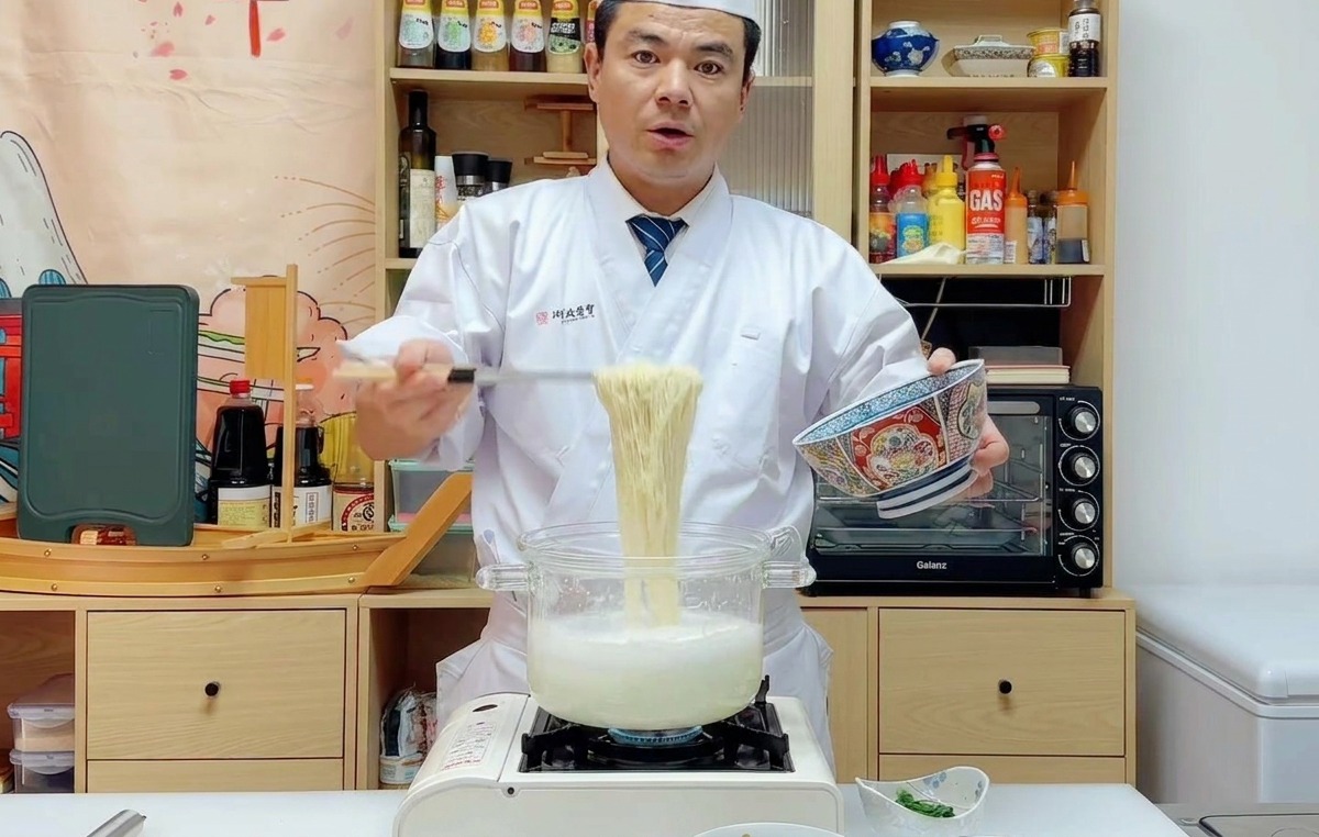 Chef Wu lifting a large portion of cooked ramen noodles with a strainer and transferring them into a blue-patterned serving bowl.