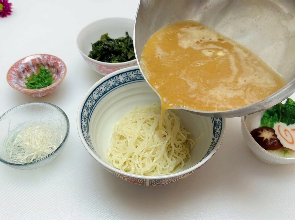 Hot, golden-brown miso soup being poured from a stainless steel pot over a mound of cooked ramen noodles in a ceramic bowl.