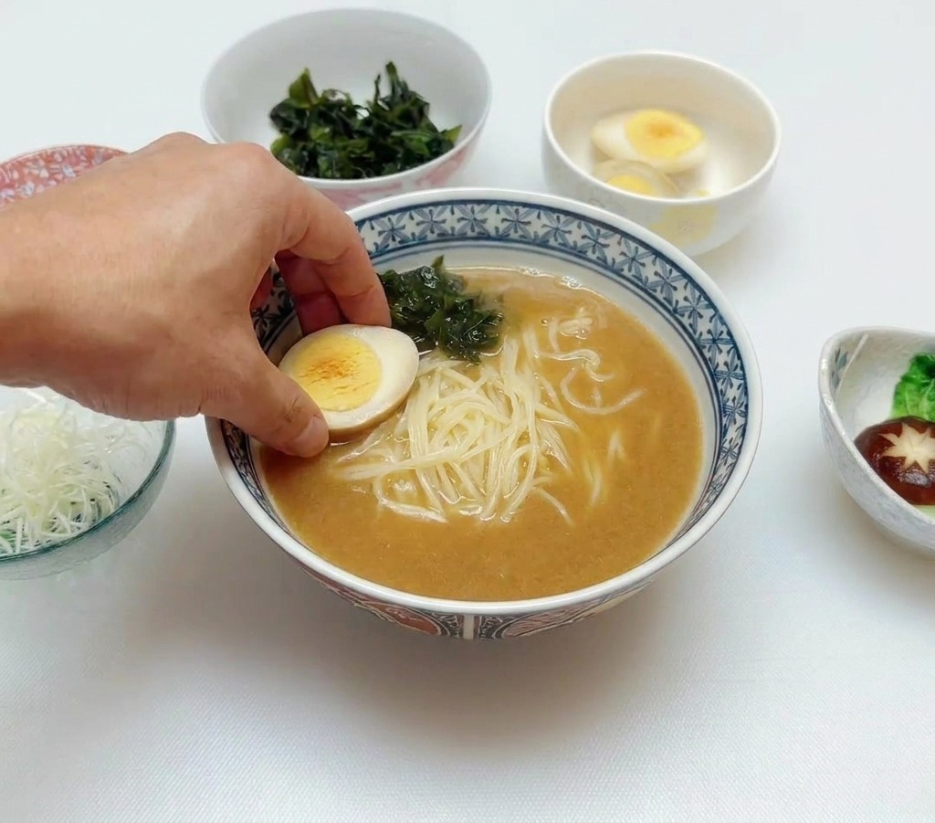 A hand placing a halved soft-boiled egg onto a bowl of ramen already containing broth, noodles, and green wakame seaweed.