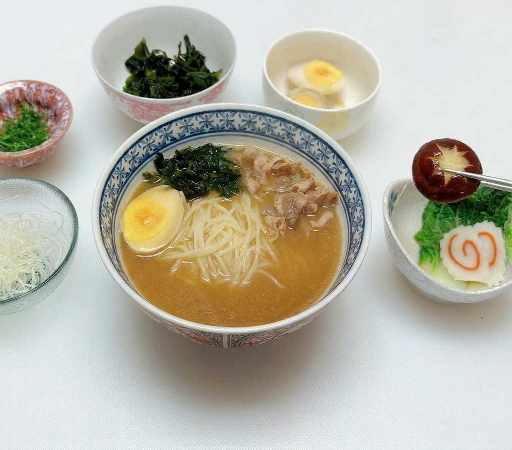 Chopsticks placing a dark brown scored shiitake mushroom onto the toppings of a miso ramen bowl that already includes an egg and fish cakes.