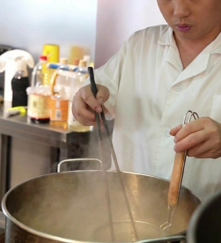 A chef lowering a wire mesh strainer filled with fresh noodles into a large pot of steaming boiling water.