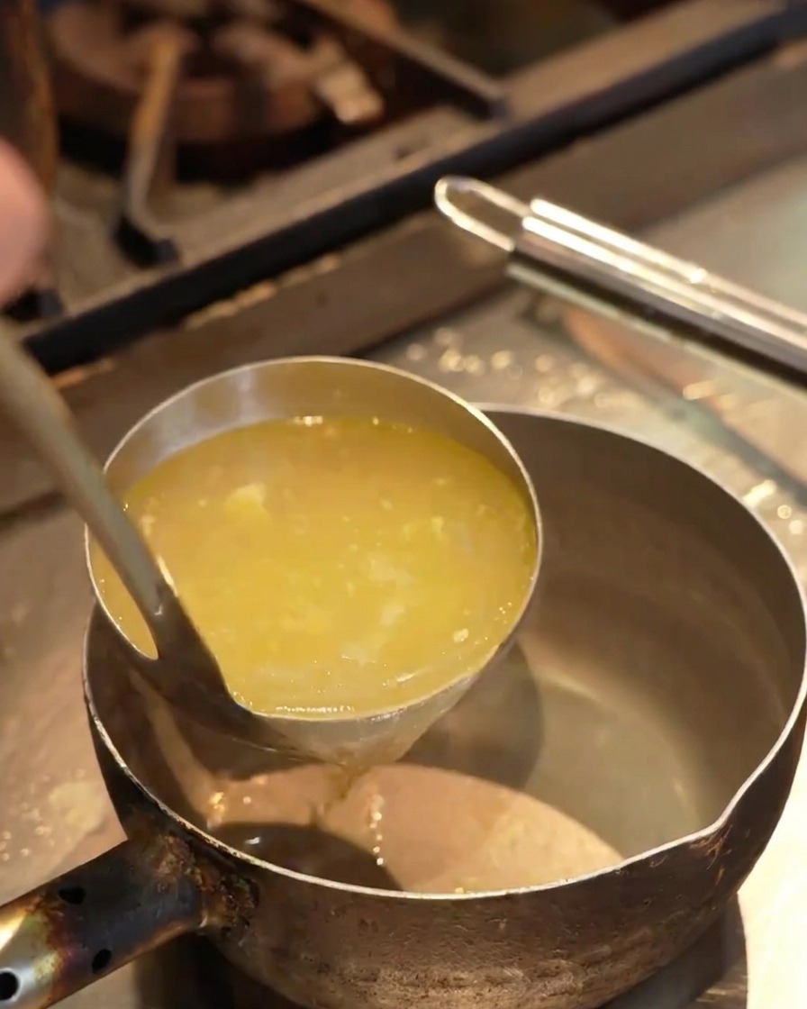 Clear yellow broth being poured from a ladle into a small handheld metal pot over a kitchen counter.