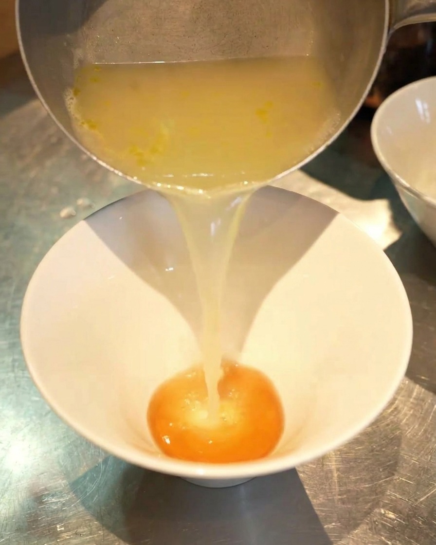 A stream of clear, golden chicken broth being poured from a silver pot into a white bowl containing a darker orange seasoning sauce.