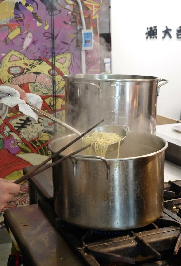 A person using a mesh strainer to lift cooked ramen noodles out of a large stainless steel pot of boiling water.
