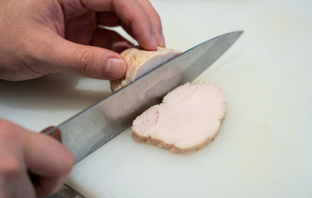 Hands of a person slicing a pale tender piece of cooked chicken breast into thin rounds on a white cutting board.