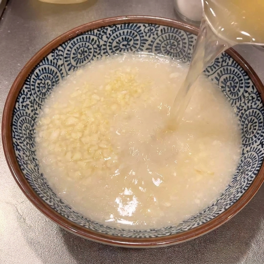 Hot, clear seasoned ramen broth being poured from a pot into a decorated blue and white ceramic bowl containing grated garlic.