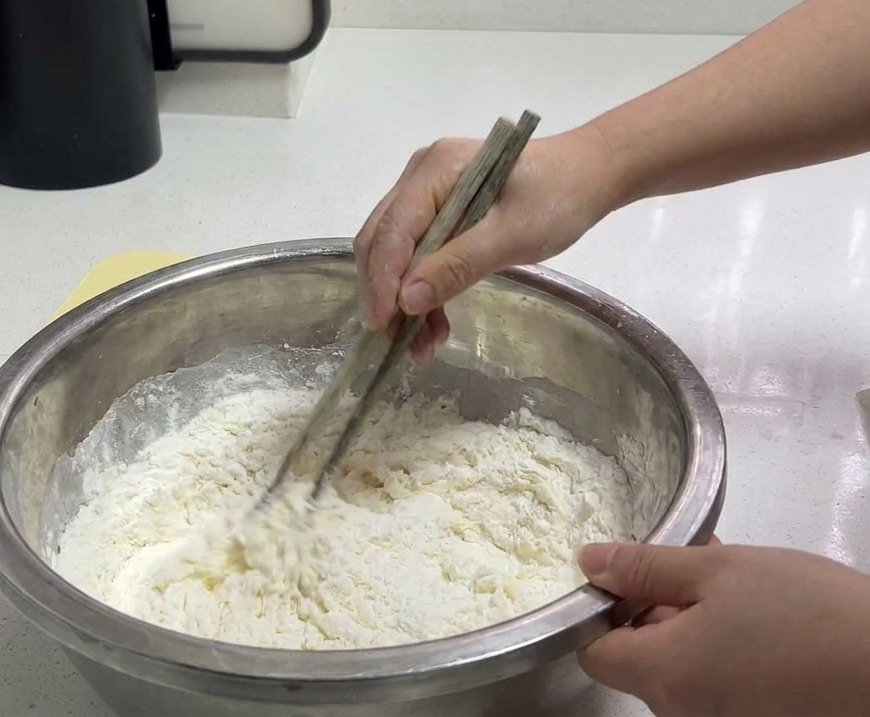 A person using wooden chopsticks to stir white wheat flour and water in a large stainless steel bowl to form a shaggy dough.