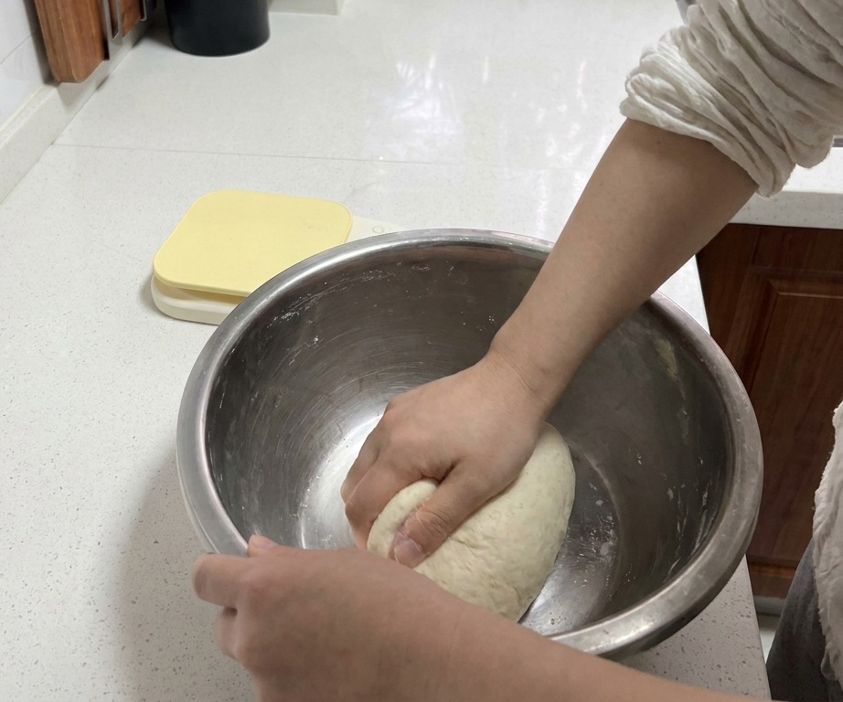 Hands firmly kneading a white dough ball inside a metal mixing bowl on a clean white countertop.