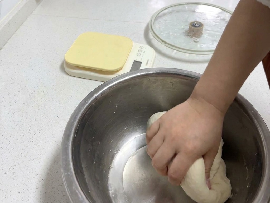 A hand kneading a smooth, round dough ball in a metal bowl to develop a delicate and elastic texture.
