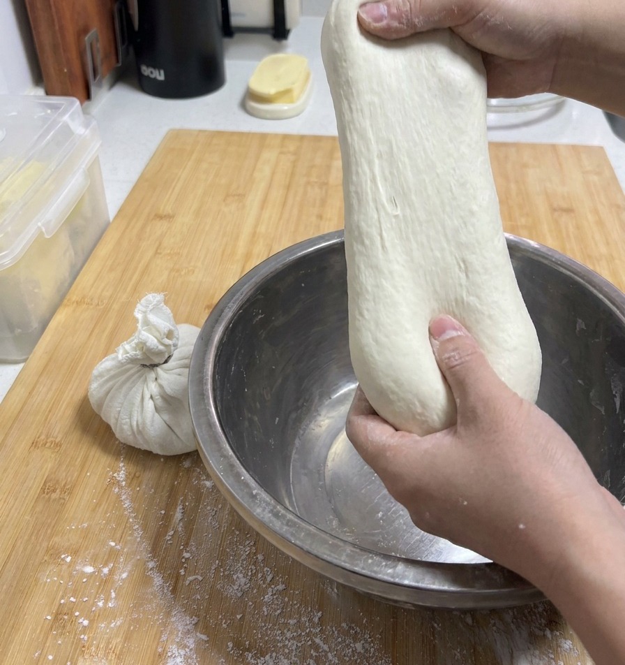 A persons hands pulling a smooth, pale piece of dough upward from a metal bowl to show its incredible stretchiness.