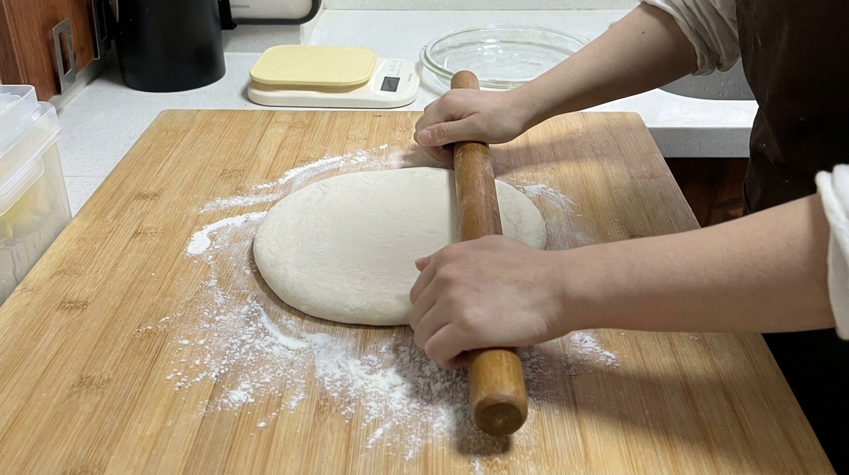 Hands using a long wooden rolling pin to flatten a round piece of dough on a bamboo cutting board sprinkled with flour.