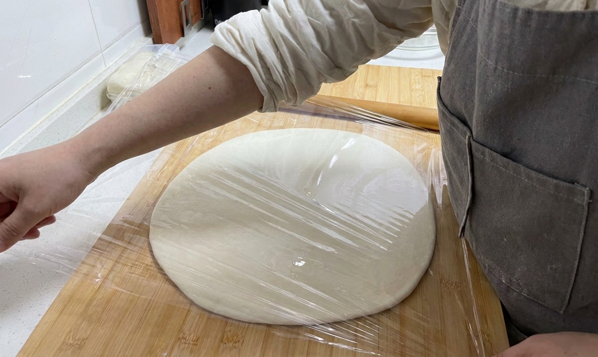A hand pulling a sheet of transparent plastic wrap over a large, flat circle of dough on a wooden board.