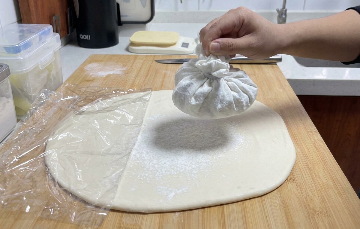 A hand holding a white cloth sachet and shaking it to release a fine mist of white flour over a flat sheet of dough.