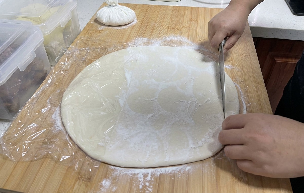 A large stainless steel knife cutting through a sheet of white dough on a wooden board to create long strips.