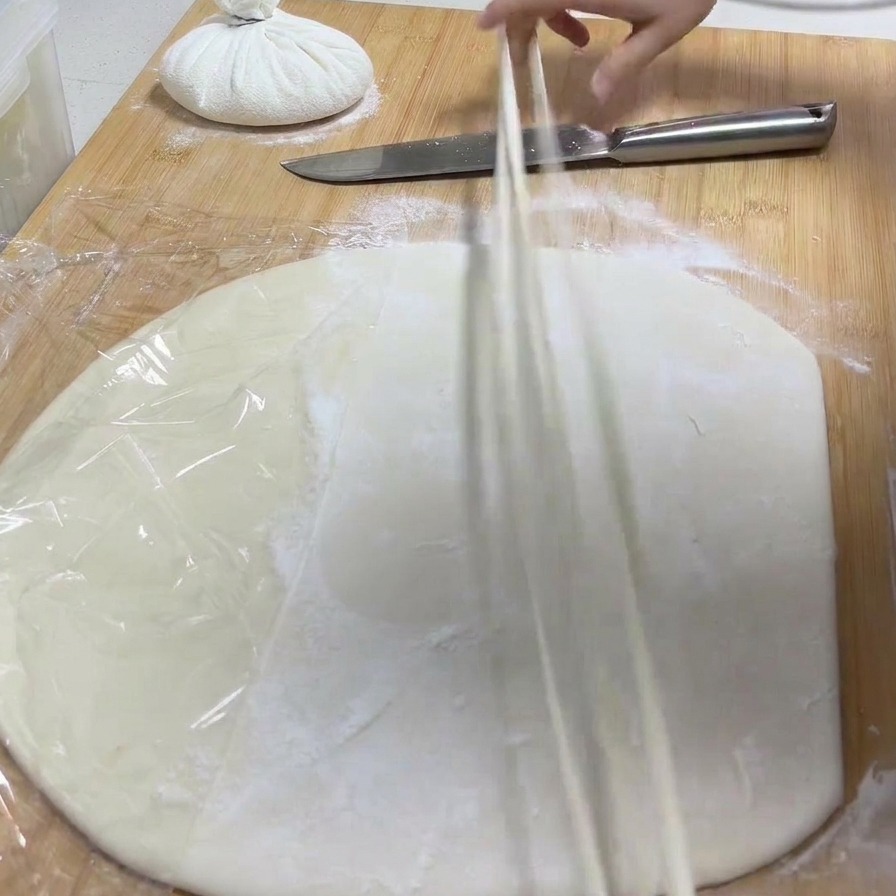 A hand lifting a long strip of dough from a board and stretching it out into a thin noodle.