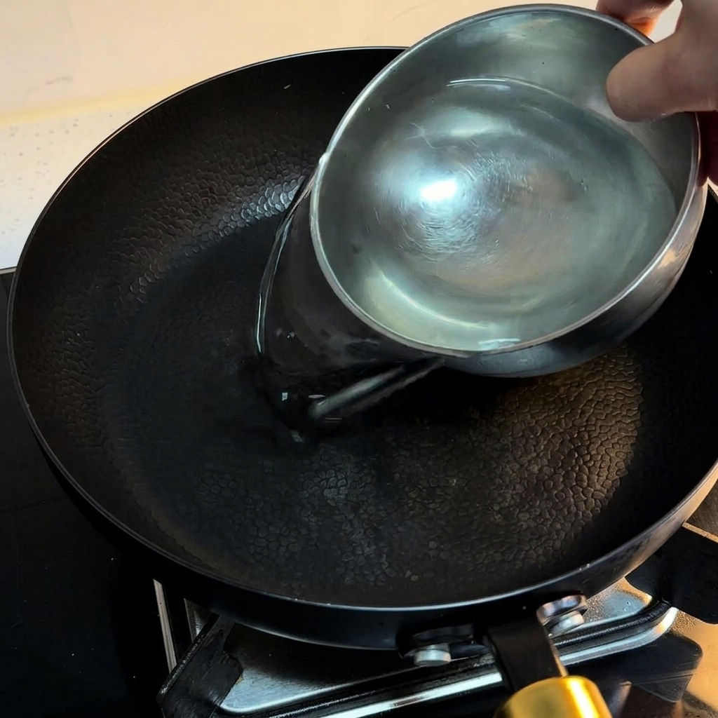 Water being poured from a small stainless steel bowl into a black non-stick pan on a kitchen stove.