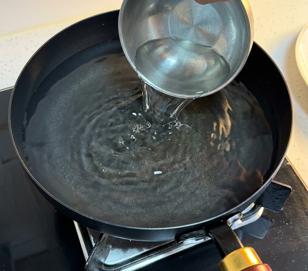 A hand pouring clear water from a small metal bowl into a black pan filled with hot water to prepare for egg poaching.